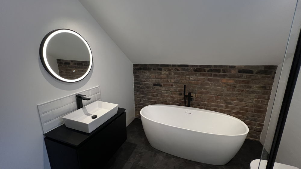 Freestanding oval bath with exposed brick-effect wall, round illuminated mirror and black vanity unit