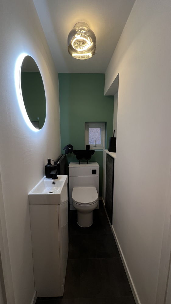 Narrow cloakroom with green wall, oval illuminated mirror, small vanity unit and toilet